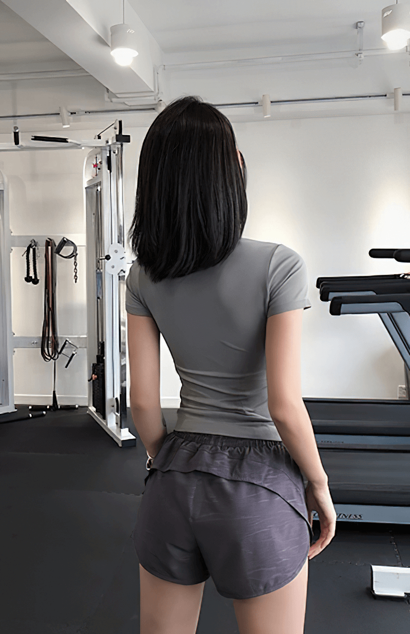 Back view of woman in gray breathable short sleeve fitness t-shirt and shorts, standing in a gym setting with equipment behind.